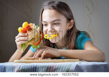 11 year old girl with pleasure eats a hamburger - a piece of bell pepper sticks out of her mouth crumbs on the table.