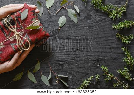 Hands holding red fabric present on a black wooden table with eucalyptus and fir tree decoration.