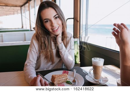 Woman sitting by the table with food on date in cafe near the sea, looking at camera and holding hand her man. First-person view