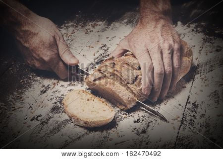 Baking and cooking concept background. Hands of baker cutting bread loaf with knife on rustic wooden table sprinkled with flour. Stained dirty hands of baker. Dark filtered