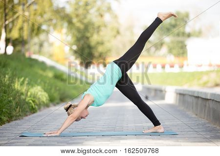 Sporty young girl practicing yoga, standing in one legged downward facing dog exercise, eka pada adho mukha svanasana pose, working out, wearing sportswear, outdoor full length, urban background