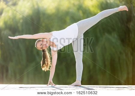 Sporty beautiful young woman practicing yoga, standing in Half Moon exercise, Ardha Chandrasana pose, working out, wearing sportswear, outdoor full length, summer street background