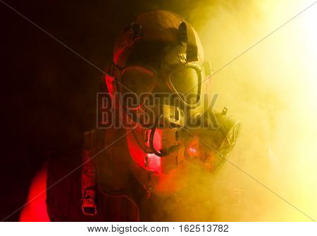A creepy guy with post apocalyptic vibes wearing a gasmask in a smoke or fog cloud that's colored bright yellow while being lit with a red light from other side.