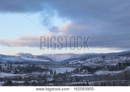 Winter landscape with clouds and clear blue sky over snow covered mountains.  Bright white mountain peaks from sunrise sunshine.  Forest trees on hills.  Houses and snow in valley in winter season.