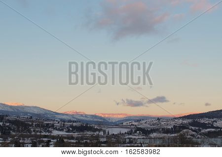 Winter landscape with clouds and clear blue sky over snow covered mountains.  Bright white mountain peaks from sunrise sunshine.  Forest trees on hills.  Houses and snow in valley in winter season.