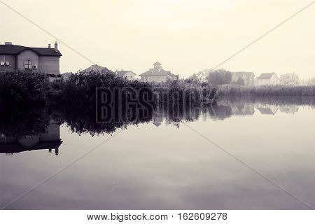 Landscape with village on bank of river. Houses reflected in water at dawn. Autumn misty morning calm. Lyrical mood. Toned black and white color processing. Nice soft warm light