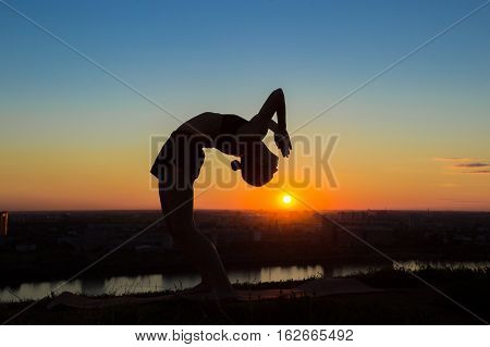 Silhouette of sporty woman practicing yoga in the park at sunset - drop back, wheel pose. Sunset light, sun lens flares, golden hour. Freedom, health and yoga concept