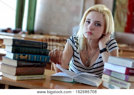 Cute blonde reading a book, sitting at a desk in a class among the piles of books. Looking into the camera