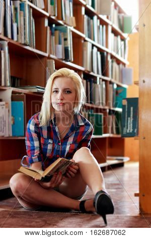 Young blonde girl in a plaid shirt with a book in hand sitting on the floor in the old library among the shelves