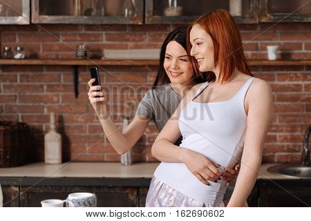 Selfie time. Happy lesbian women standing in pajamas in semi position looking at mobile phone while making selfie