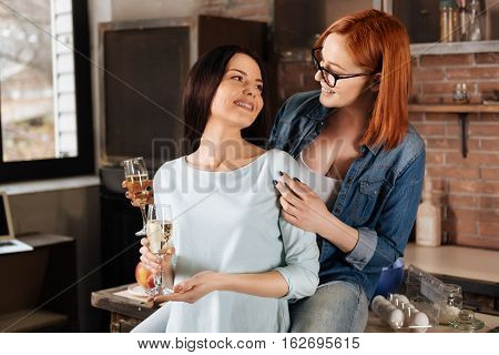 Good in the air. Red haired woman holding a glass of fizzy drink while sitting on the table looking and touching shoulder of her friend