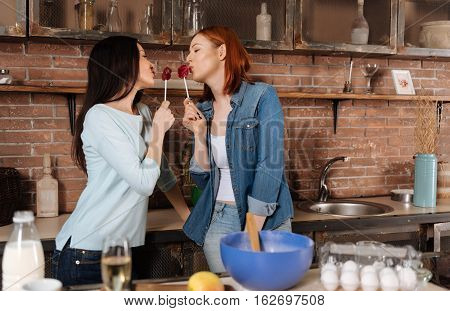 Sweet candies. Cute delighted women wearing casual clothes holding candies in front of lips standing in semi position in kitchen