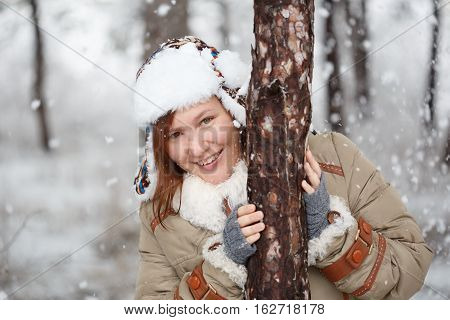 Young beautiful smiling woman in coat with white fur hat with ear flaps and grey fingerless knitted mittens hugs a tree in winter forest with falling snow around. Shallow dof. Focus on eyes.
