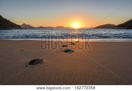 Beautiful sunrise with the sun rising from the Atlantic Ocean, solar path on the water and footprints at the deserted Praia Vermelha Beach, Rio de Janeiro, Brazil