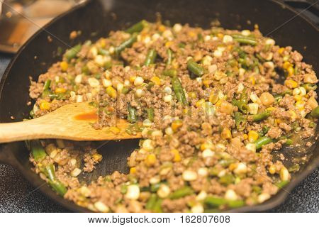 Cooking a casserole with ground meat, green beans, corn, Home made casserole in an iron skillet with wooden spatula.