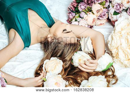 Sexy young pretty woman or cute girl with long hair in green dress laying on white bedsheet among lilac rose and peony flowers on wooden floor