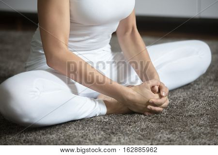 Close up of a young woman practicing yoga, sitting in Butterfly exercise, baddha konasana pose, working out, wearing white sportswear, indoor, home interior background