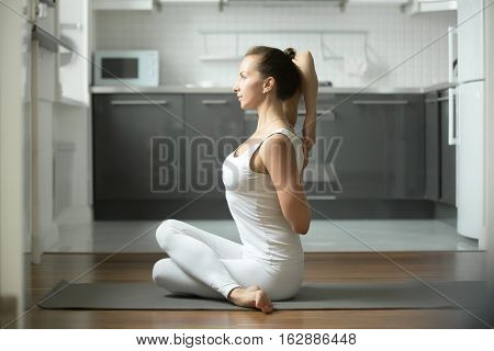 Sporty young attractive woman practicing yoga, sitting in Cow Face exercise, Gomukasana pose, working out, wearing white sportswear, indoor full length, home interior background