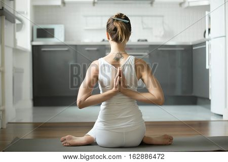 Sporty attractive woman practicing yoga, sitting in variation of Cow Face exercise, Gomukasana pose, working out, hands in namaste behind the back, indoor, home interior background. Rear view