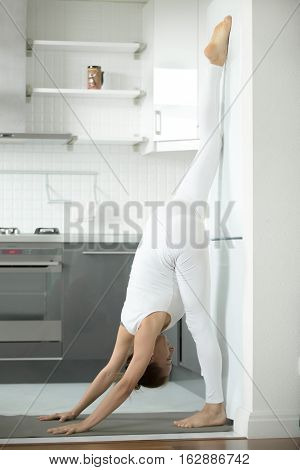 Sporty attractive woman practicing yoga, standing in one legged downward facing dog exercise, eka pada  adho mukha svanasana pose at fridge, working out, indoor full length, home interior background