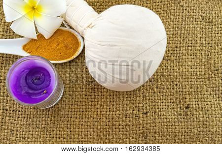 Spa herbal compressing ball white frangipani flowers (Plumeria spp Apocynaceae Pagoda tree Temple tree) turmeric powder in wooden spoon and candle on brown sack fabric background