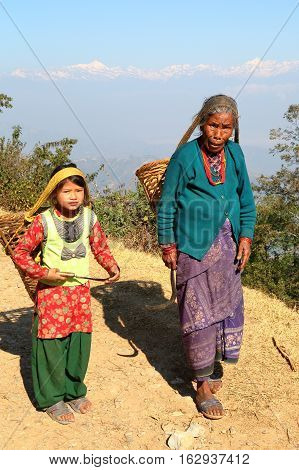 DHULIKHEL, NEPAL - DECEMBER 25, 2014: An old woman and a little girl walking in the countryside with a basket on their back (himalayan mountains in the background)