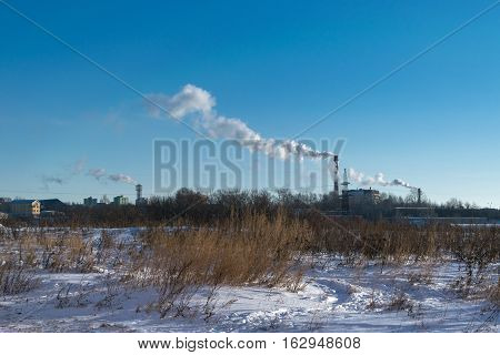On the background of snow-covered vacant lot visible pipe of a thermal power plant with large pipes. Of the pipes coming out thick smoke polluting the atmosphere.