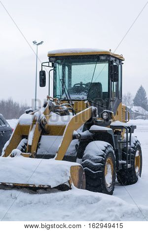 yellow wheeled snow plow tractor with a bucket all covered with snow stands on the cleared the Parking lot after a heavy snowfall