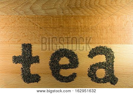 The word tea is laid out on a light-brown wooden Board small leaf black tea. Board is the background for the word tea.