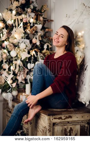 Woman with a Christmas Gift sitting on acncient commode decorated for xmas. Fir tree at background.