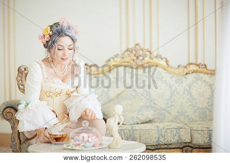 Greyhead Woman In Rococo Dress Posing In Front Of Pink Background While Drinking Tea