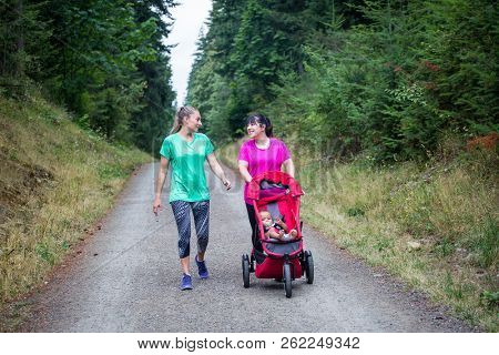 Two women in their 30s walking and talking together on a trail with pushing a baby stroller