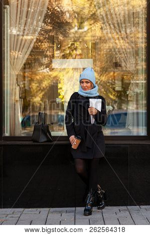 Young Attractive Lady In Blue Knitting Hat And Black Coat Drink Coffee To Go . Smiling Beautiful Hip