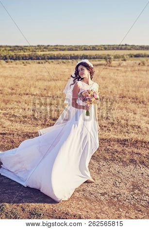 Beautiful Smiling Bride Wearing A Natural Flower Wreath And Standing In The Golden Summer Fields Wit