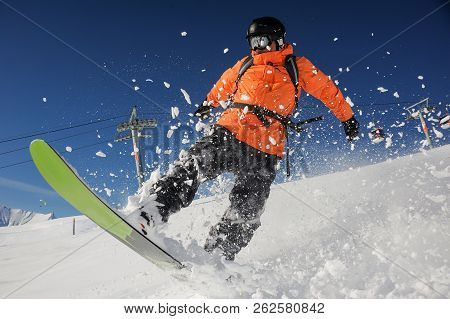 Snowboarder Dressed In Orange Sportswear Riding Down The Powder Mountain Hill On Sunny Day. Snowboar