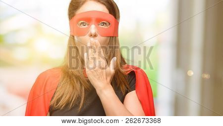 Middle age super hero woman wearing red mask and cape covers mouth in shock, looks shy, expressing silence and mistake concepts, scared