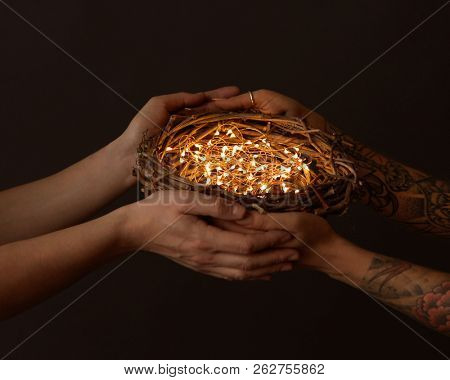 A nest of branches with bright garlands are held by male and female hands with a tattoo on a black background. Celebration