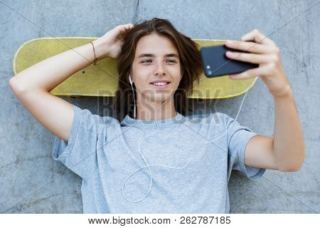 Top view of a smiling young teenge boy spending time at the skate park, laying on a skateboard, taking a selfie, listening to music with earphones