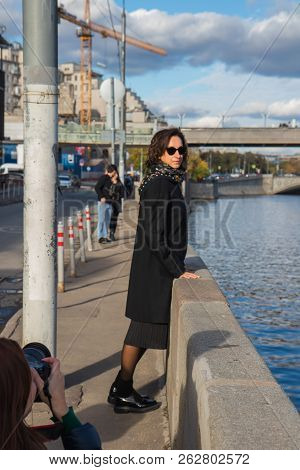 Photographer In The Street Photo Shooting Process. Model Is Young Lady In Stylish Black Coat With Bl