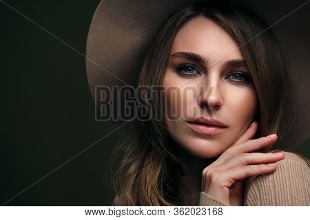 A Young Attractive Blonde Woman In A Beige Sweater, Fedora Hat And Brown Leather Trousers Poses On A