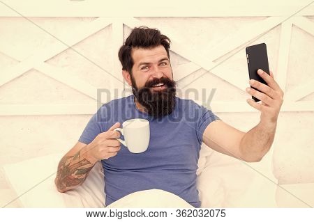 Source Of Energy. Bearded Man Enjoy Video Conferencing And Coffee In Bed. Happy Hipster Hold Cup And