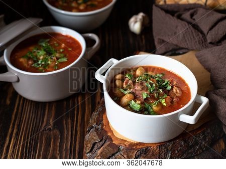 Bean Soup With Meat And Vegetables Served On A Rustic Board And Wooden Table With Bread And Garlic. 