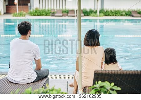 Young Family Resting By Swimming Pool And Looking At Clean Turquoise Water, View From The Back