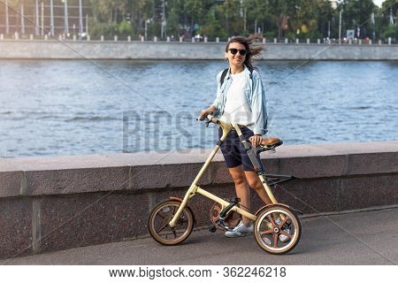 Beautiful Happy White Woman In Sunglasses Stands On The Embankment Of A Wide River. In The Hands Of 