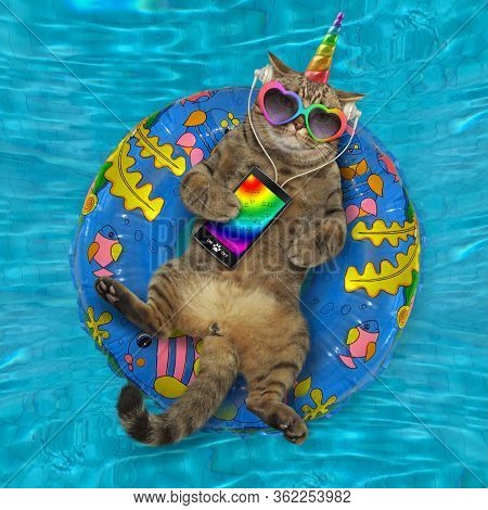 The Beige Cat Unicorn In Headphones And Sunglasses With A Smartphone Is Lying On An Inflatable Blue 