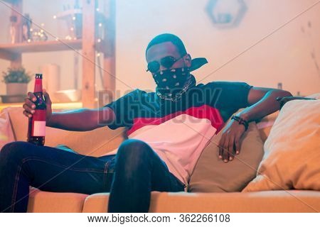 Young African man in black bandana on face, jeans and t-shirt having bottle of beer while relaxing on soft couch at home