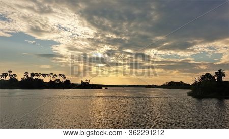 Sunset On The Zambezi River. The Expanse Of The River, Low Clouds, Tree Silhouettes And The Sun Sink