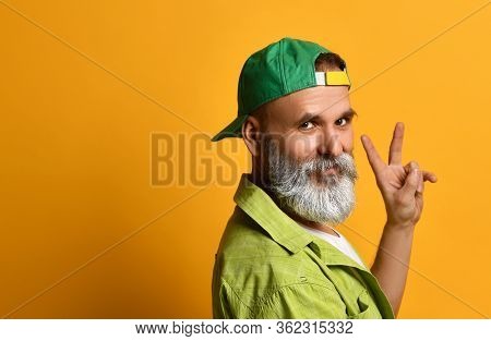 Gray-bearded Man In A Green Cap And Shirt, A White T-shirt, A Bracelet. Smiling, Showing Fingers Pea