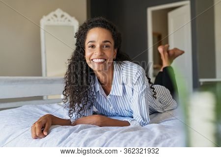 Cheerful young multiethnic woman looking at camera and smiling while lying on bed. Pretty casual mixed race woman lying on front on her bedroom. Portrait of beautiful african american girl resting.