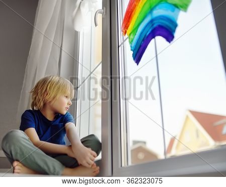Little Boy Sitting At Window With Drawing Rainbow While Coronavirus Quarantine. Rainbow Sign Is Symb